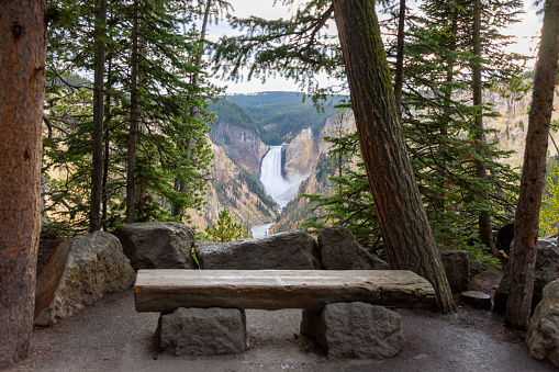 A spectacular and dramatic view of the Upper Falls, a 109-foot (33 m) waterfall on the Yellowstone River in Yellowstone National Park, Wyoming and the bench view point.  The waterfall is seen from Artist Point a platform that offers a dramatic view of the surging water. The sky above is a mix of dark rainy clouds and bright orange hues, as the sun peeks through the gaps, creating a stunning contrast with the green trees and the brown rocks.