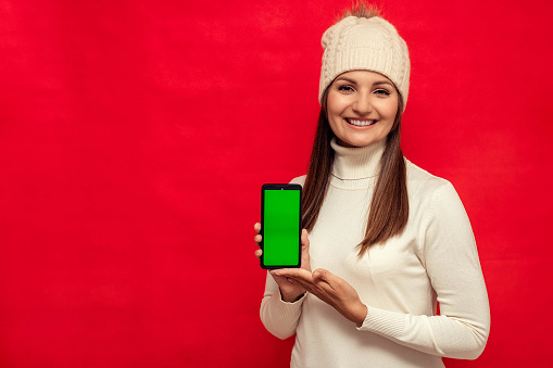 Young woman in a white jumper and a white hat holds a smartphone with a green screen on a red background, copy space, chroma key.