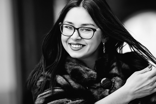 Close-up face of a girl in glasses and winter clothes that looks at the camera lens and smiles on black and white photography