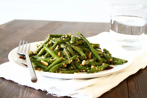 Glazed green bean salad with crushed pine nuts on a white plate with a fork.