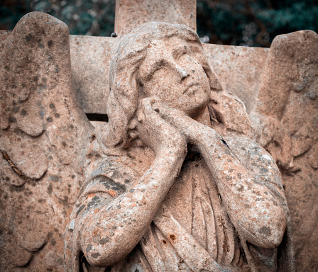 A contemplative and rather soulful reconstituted stone angel on a cross in a suburban graveyard.