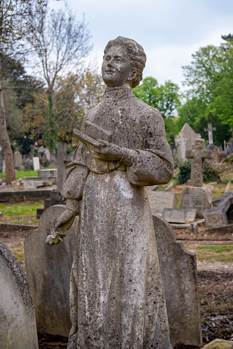 A generic figure of a woman holding a prayer book or bible in her left hand, plus a rose in her right hand, on a grave in a suburban cemetery. Such figures were very popular during the late 19th and early 20th centuries and were made of reconstituted and moulded stone. This example has survived without much damage except natural weathering.