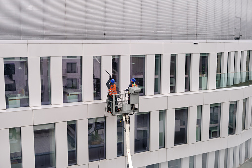 Wroclaw, Poland - Apr 14, 2021. Two workers wearing safety harness wash walls, panels and windows of office building facade at height standing in a crane cradle or aerial platform using pressure washer, wipers and mops. Daytime, Europe, cleaning facades, modern architecture.
