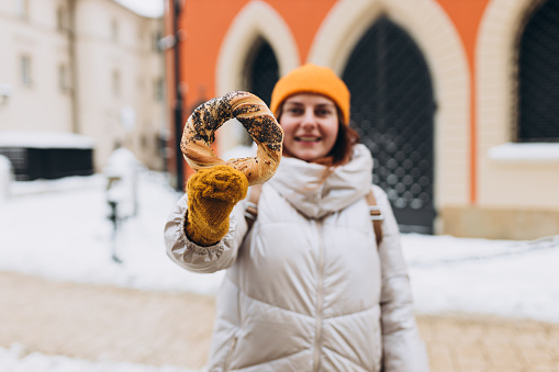 Attractive young female tourist in yellow hat is holding pretzel, traditional polish bagel on Market Square in Krakow. Traveling Europe in winter time.