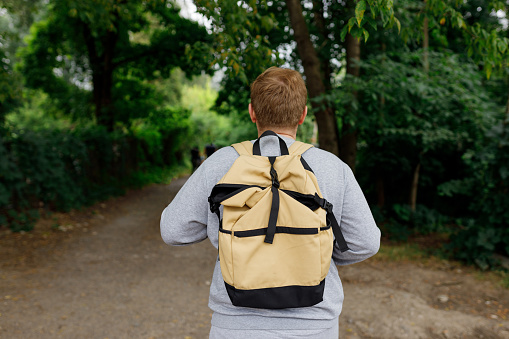 Rear view of man with backpack hiking in forest or city park. Man in active trekking clothes walking in the countryside, back view