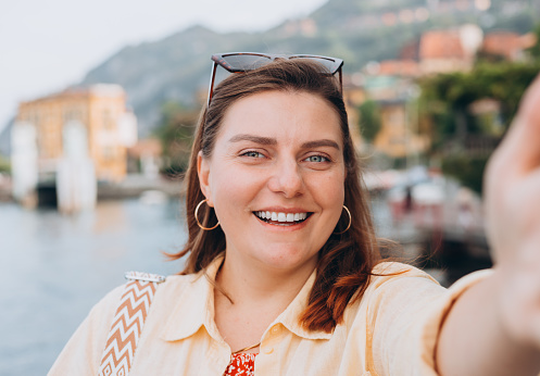 Photo relaxed charming positive young woman hold camera and make selfie summer outdoors. Concept of vacation and travelling. Varenna, Italy, Como
