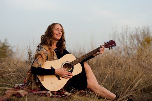 beautiful young smiling woman playing melody on acoustic guitar while sitting in field among tall dried grass. Talented musician. Autumn sunset. Outdoor picnic. Romantic mood. Creativity, lifestyle