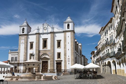 This renaissance church dedicated to Saint Anthony the Great overlooks the main square (Praca de Giraldo) in the Portuguese city of Evora. A beautiful sunny day in late summer with blue sky and some light clouds. Incidental people are having lunch shaded by the sun umbrellas of on outdoor restaurant in the square.