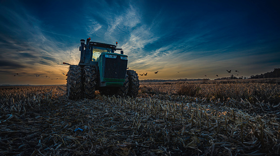 Witness the breathtaking beauty of this captivating moment as the sun sets behind the horizon, casting a warm and dramatic glow across the expansive fields. Our striking photograph features a powerful tractor, silhouetted against the vibrant hues of the twilight sky. The interplay of light and shadows adds a touch of cinematic flair, making this image a perfect choice for conveying both the tranquility and power of rural life at dusk. Download this evocative shot now to infuse your projects with the essence of rural charm and cinematic drama.