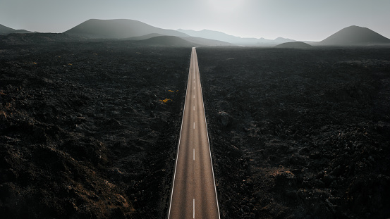Endless straight highway towards the horizon through the Timanfaya National Park Black Volcanic Landscape. Shot against sunset light and Volcano Mountains on the Horizon. Drone Point of View Stitched Panorama DJI Mavic 3 Pro. Timanfaya National Park, Lanzarote Island, Canary Islands, Spain - Africa.