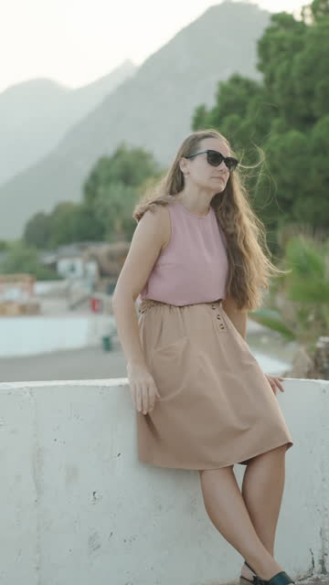 Vertical video. A Young Woman leans against a concrete wall with a backdrop of green hills and trees. A beach by the sea. In slow motion.