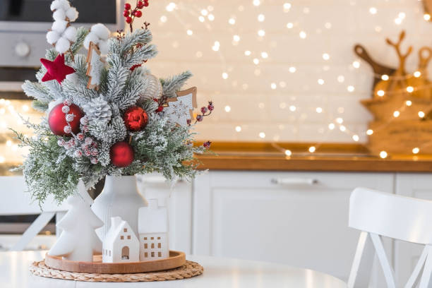 A spruce bouquet with red berries, toys, cotton on a white kitchen table against a beautiful garland. Christmas decorations in the interior. The concept of preparation for the New Year holidays 2024.