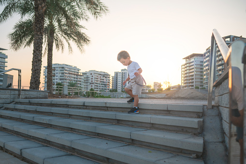 Cute multiracial toddler in a summer outfit is walking down the stairs outside. In the background are buildings and a palm tree