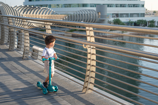 Cute multiracial toddler in a summer outfit is having fun while riding a blue children's three wheeled scooter outdoors, near the sea. Children's sports, vehicle for active children, free activity for boys and girls