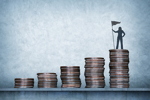 A silhouette of a woman standing on the tallest stack of a row of coin stacks plants her flag as she has reached her financial target.