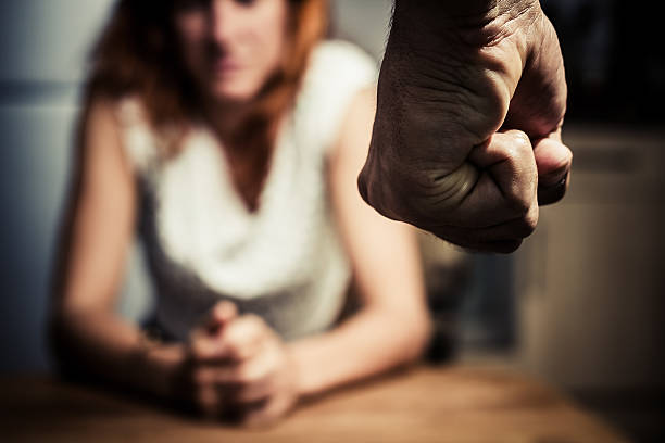 Woman in fear of domestic abuse Young woman is sitting hunched at a table at home, the focus is on a man's fist in the foregound of the image wife stock pictures, royalty-free photos & images
