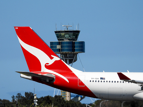 A Qantas Airbus A330-202 plane, registration VH-EBR, taxis past the air traffic control tower of Sydney Kingsford-Smith Airport as preparation for departure as flight QF83 to Singapore. The airport' security fence and surveillance cameras are visible in the bottom left. This image was taken from Botany Bay, Kyeemagh on a sunny afternoon on 18 November 2023.