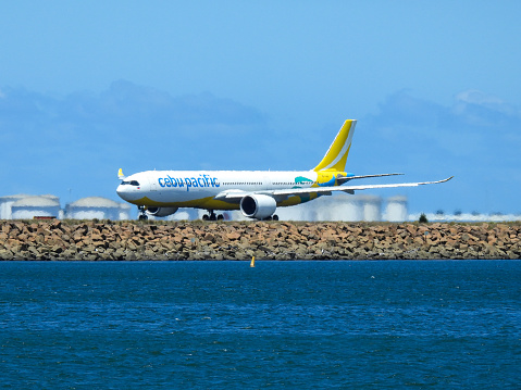 A Cebu Pacific Airbus A330-941 plane, registration RP-C3902, powering down the main north-south runway of Sydney Kingsford-Smith Airport as flight 5J39 to Manila.  In the background are fuel storage tanks.  A yellow buoy marks the airport exclusion zone. This image was taken from Botany Bay, Kyeemagh on a sunny afternoon on 18 November 2023.
