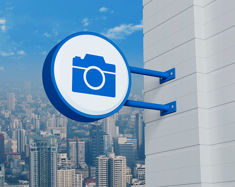 Camera icon on hanging blue rounded signboard over modern city tower, office building and skyscraper, Business camera service concept, 3D rendering