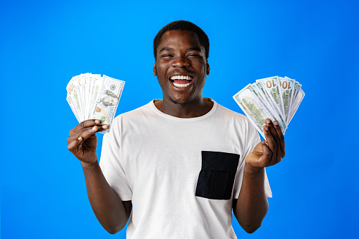 Young african man holding cash money in dollar banknotes against blue background close up