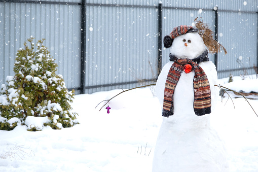 Funny snowman in a hat and scarf on the background of grey fence in the yard. Winter, winter entertainment, snowfall