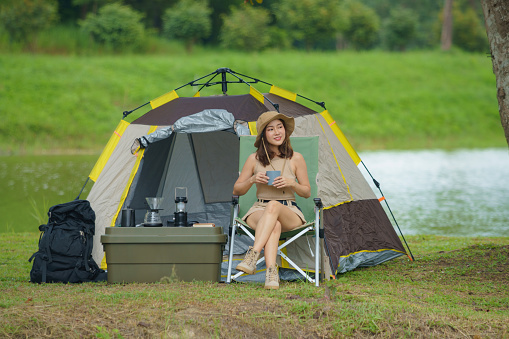 An Asian woman is enjoying sitting near a lake and drinking hot coffee in nature while in a tent camp outdoors camping.