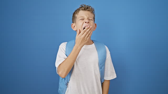 Adorable blond boy student, exhausted from studying, stretching arms back in a tired yawn against isolated blue background