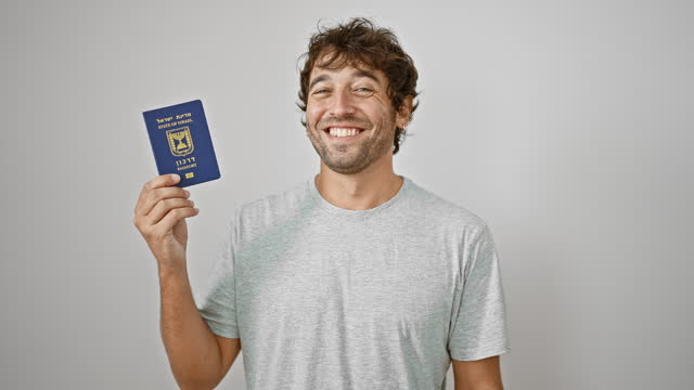 Joyful young man, casually sitting on a white sofa, flashing his israeli passport with a confident smile against an isolated background