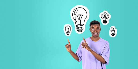 Portrait of excited young African man college student pointing at light bulb drawings standing over green copy space background. Concept of bright idea and brainstorming