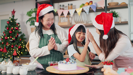Happy Grandmother, mother and little daughter decorated cake together for Christmas Eve. Three generations Asian women making cake for Christmas day. Christmas cake decorated. Festive holiday