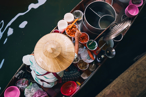 Selective focus on traditional hat of vendor on boat on floating market. Woman is preparing food on water in Damnoen Saduak near Bangkok, Thailand.