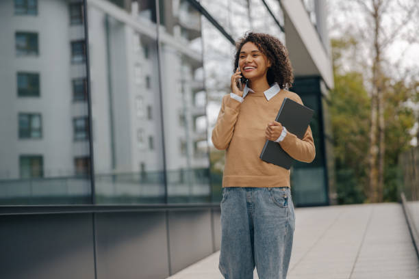 istock Pretty female entrepreneur is talking phone while standing with laptop on modern building background 1802035662