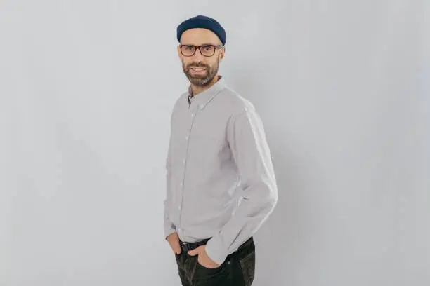 Profile shot of attractive unshaven man dressed formally, wears optical glasses, models over white background, happy to achieve good results at work, has pleased facial expression. People and style