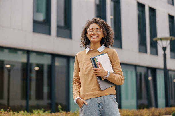 istock Young smiling female student standing with laptop and note pads on modern building backgrounds 1802059462