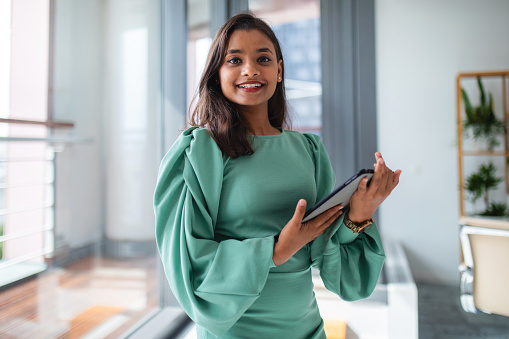 Professional Indian woman smiling at the camera while standing in a bright cosy office. She is looking at the camera while holding a digital tablet in her hands. The woman looks happy and confident. Another day at the office as a female CEO.