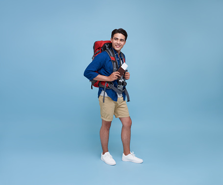 Happy young Asian tourist man holding passport with backpack going to travel on holidays isolated on blue background.