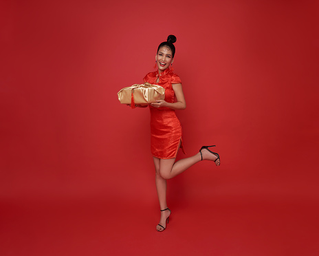 Happy smiling Asian woman in red traditional costume giving gold gift box for bless chinese new year isolated on red background.