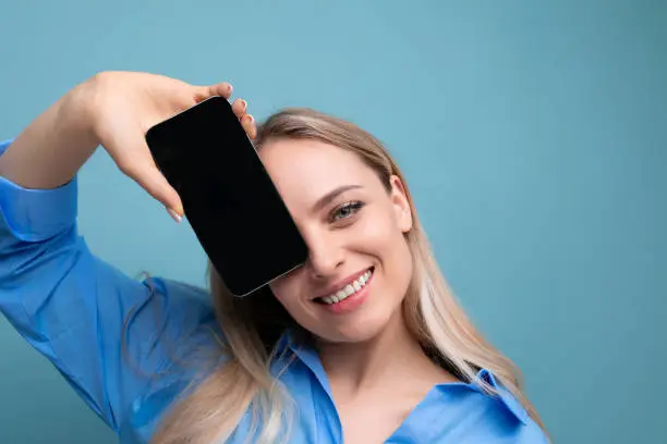 Photo of smiling stylish blond girl holding smartphone screen with mockup for web page near her face on blue background