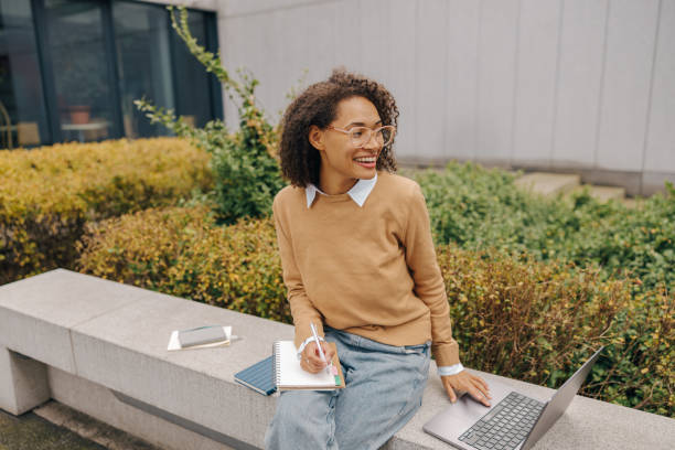 Smiling business woman working laptop and making notes sitting on background of office building