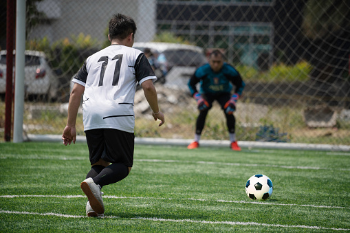 Male soccer athlete prepares to kick a penalty shot on the field while his teammates watch. The opposing goalie is protecting the goal. Photo is shot from the rear view. The player has his leg behind him while preparing to kick the ball