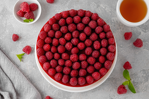 No baked raspberry cheesecake or raspberry cream mousse cake with jelly and fresh berries on top on a white plate on a gray concrete background. Summer cake