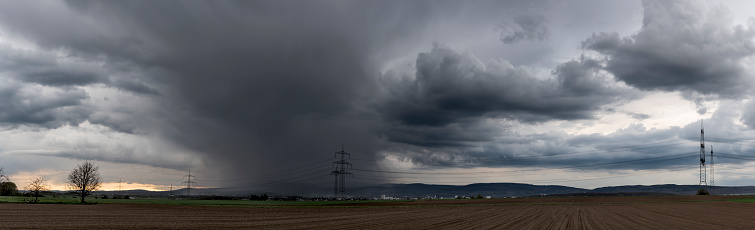 Panoramic view of a thunderstorm moving over the Taunus with grey clouds, with ploughed and fallow fields in the foreground and a high-voltage overhead power line