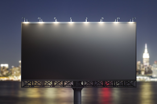 Blank black billboard on city buildings background at night, front view. Mockup, advertising concept