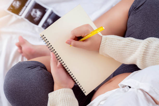 nabiał ciążowy kobieta w ciąży zapisuje harmonogram w notatniku na pamięć matka siedzi na łóżku z ultrasonografią dziecka kobieta w ciąży sporządza listę rzeczy do spakowania do szpitala położniczego widok z góry plan matki dla dziecka - human pregnancy telephone ultrasound family zdjęcia i obrazy z banku zdjęć