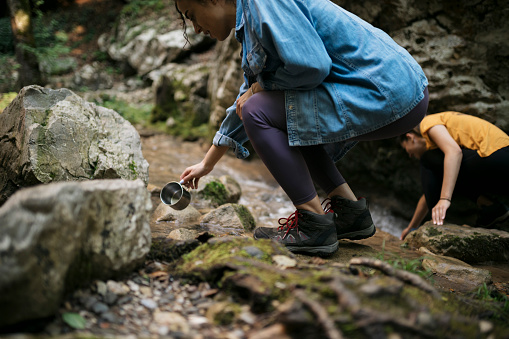 Women hikers washing coffee cups in mountain stream on hiking trip. Female tourists washing utensil in mountains river while on forest hike.