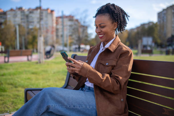 istock Smiling beautiful woman text messaging while sitting on a park bench on sunny day 1802245321