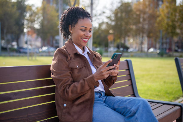 istock Smiling beautiful woman using phone while sitting on park bench on sunny day 1802246401
