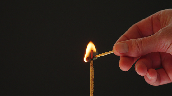 Close-up of human hand holding match and incense stick against black background. Photo taken November 19th, 2023, Zurich, Switzerland.