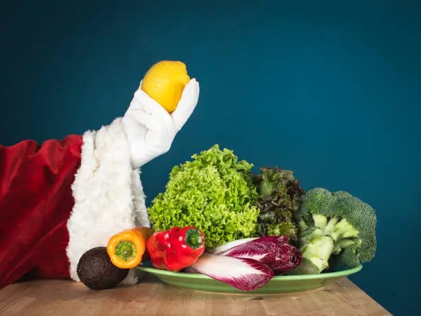 Santa's hand holds a yellow citron. There are red vegetables on the table: on a plate are sweet paprika, broccoli, lettuce, lettuce and endive. On a turquoise background is space for the text.