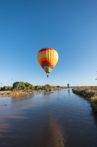 A vibrant multicolor hot air balloon flies low over the Rio Grande River during the Albuquerque International Balloon Fiesta, its reflection visible in the surface of the water. More balloons can be seen further upriver in the distance.